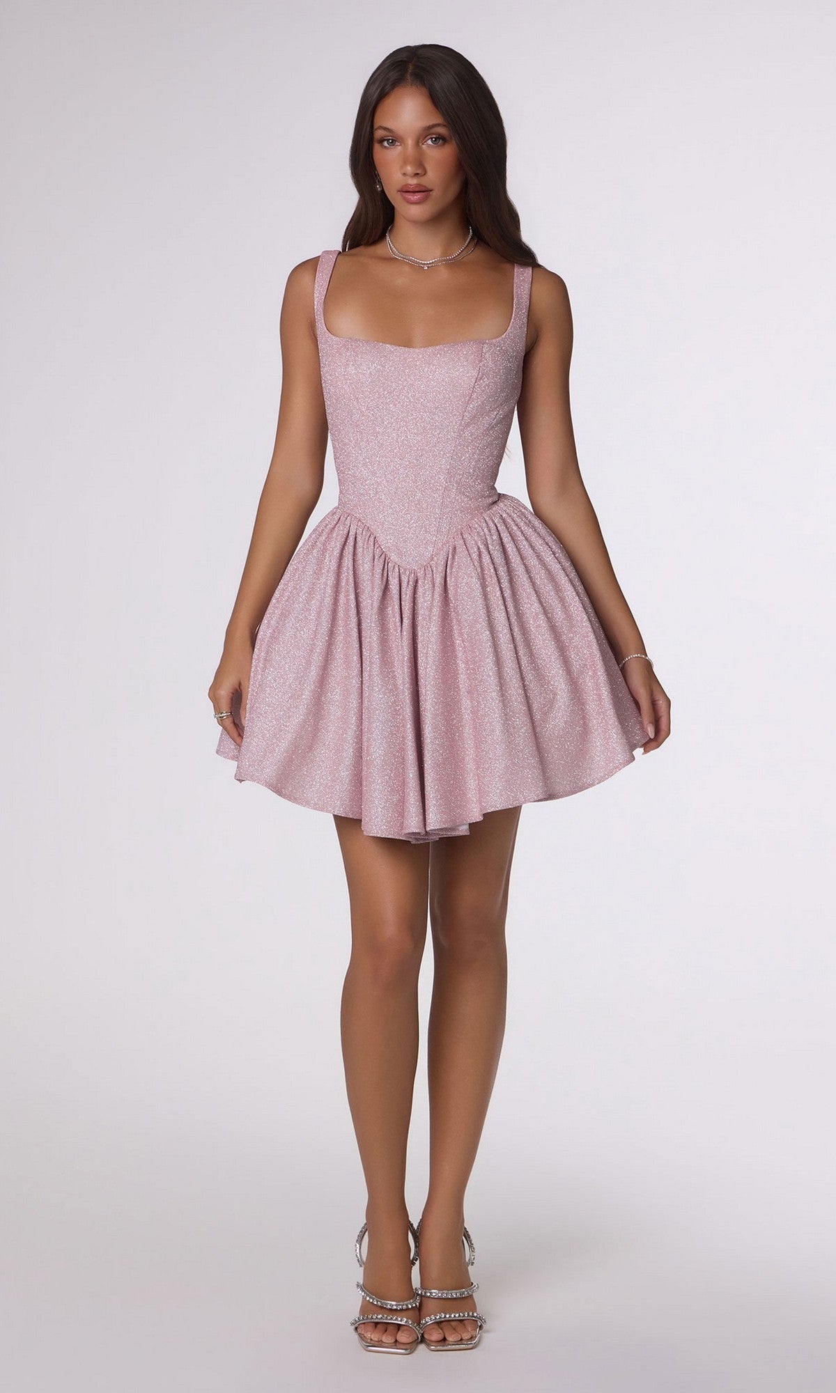 A woman poses against a plain background wearing the Short Homecoming Dress: Amoris T738—a light pink, fit-and-flare mini dress made from shimmering stretch fabric—paired with silver high-heeled sandals.